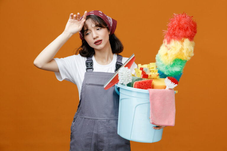 tired young female cleaner wearing uniform and bandana holding bucket of cleaning tools touching head looking down isolated on orange background